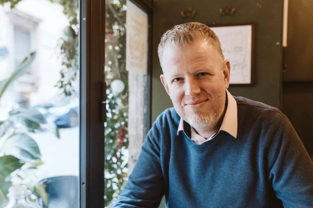 Herr Herscheid mit blauem Pullover sitzt an einem Tisch vor einem Fenster mit Pflanzen im Hintergrund